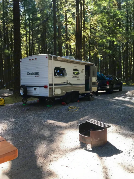 Dutchmen Sport RV rental set up in a sunny forest campsite with a pickup truck and fire pit for outdoor fun.. Dutchmen Sport 2004