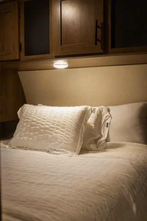 RV rental bedroom: cozy bed with white bedding, pillows, overhead cabinets, and warm lighting.. Avenger ATI 2020
