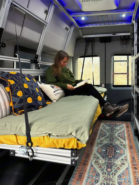 Cozy RV rental interior: A woman reads on a modular bed in a camper van, featuring comfy pillows & a stylish rug.. Vandoit DO 2019