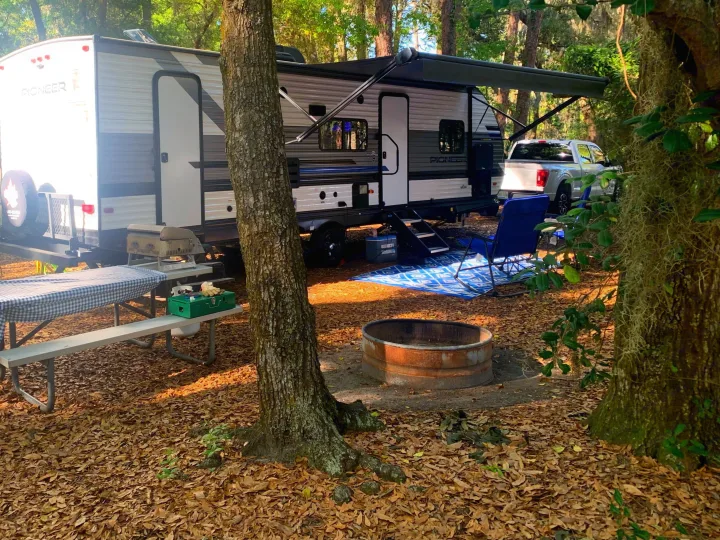 RV rental campsite with a travel trailer, white truck, picnic table, grill, fire pit, and chairs in a wooded area.. Heartland Pioneer 2022