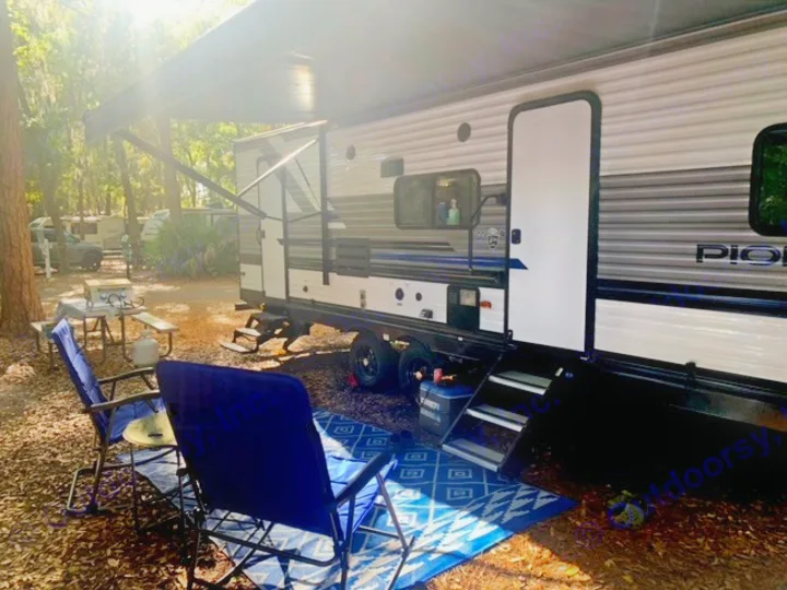 This RV rental offers a relaxing campsite with an extended awning, outdoor chairs, a blue rug, and a picnic table with a grill.. Heartland Pioneer 2022