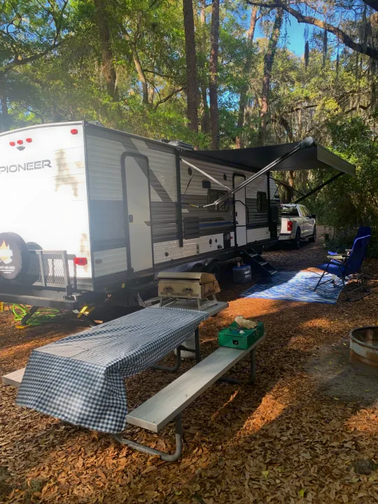 RV rental campsite featuring a Pioneer travel trailer, truck, picnic table with grill, chairs, and fire pit in a sunny forest.. Heartland Pioneer 2022