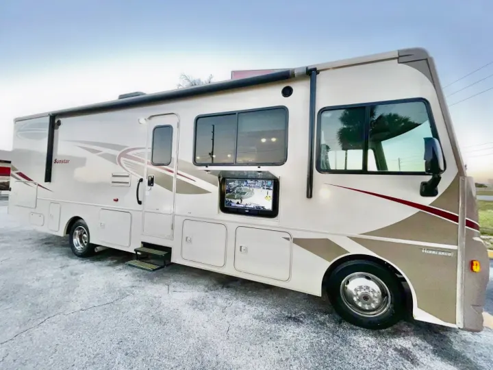 Beige and brown Winnebago Sunstar rv rental with an open outdoor TV and side awning, parked on pavement.. Winnebago Sunstar 2016