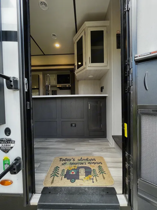 View from the open door into an RV rental, showcasing a modern kitchen with dark cabinets, wood-look flooring, and a decorative welcome mat.. Keystone Sprinter 2023