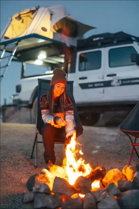 Cozy camping night with an RV rental! A woman smiles by a campfire, hands near flames. A white Jeep Gladiator & rooftop tent behind.. Jeep Gladiator 2022