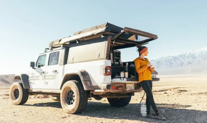This RV rental Jeep Gladiator is fully equipped for overlanding. A woman enjoys the built-in kitchen in a desert landscape.. Jeep Gladiator 2022