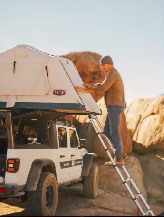 A man on a ladder adjusts a Tepui rooftop tent mounted on a white Jeep Gladiator, part of an RV rental for off-road adventures.. Jeep Gladiator 2022