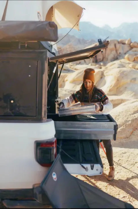 Woman organizes her RV rental's overland kitchen from the truck bed. Featuring a rooftop tent, she prepares for desert camping.. Jeep Gladiator 2022