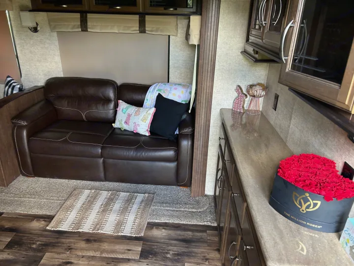 Inside an RV rental: a brown leather sofa on the left, with decorative pillows and a rug. Right side shows kitchen counter with roses.. Forest River Sandpiper 2018