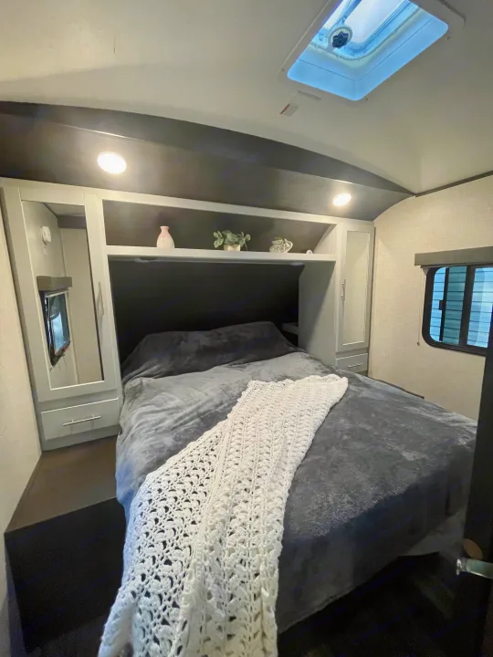 Full view of a modern RV rental bedroom featuring a comfy grey bed, white crochet throw, overhead shelving, and side window.. Keystone RV Bullet Premier Ultra 2020