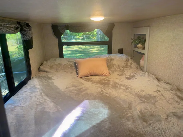 Inviting RV rental bedroom with a large, plush bed, pink pillow, and sunny windows overlooking green outdoor space.. Keystone RV Bullet Premier Ultra 2020