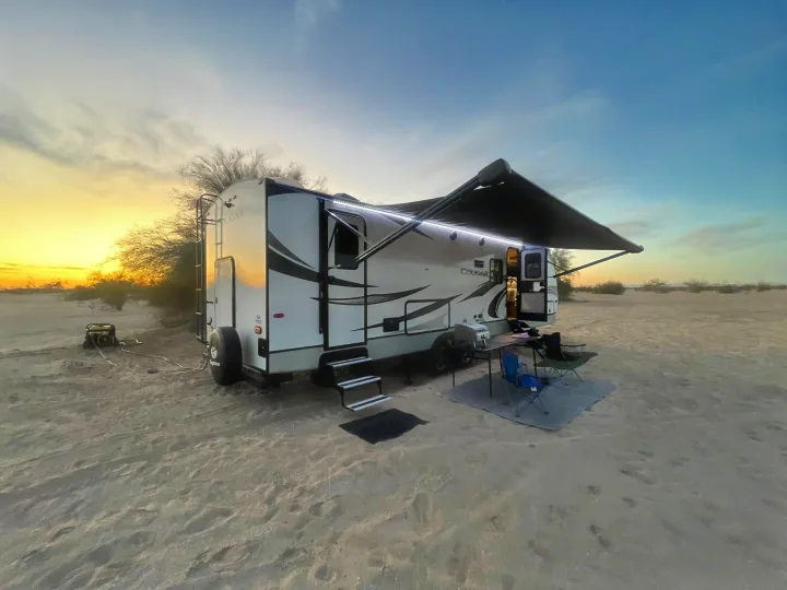 Sunset desert camping with an RV rental setup. The travel trailer features an illuminated awning, outdoor chairs, and a generator.. Keystone RV Cougar 2021