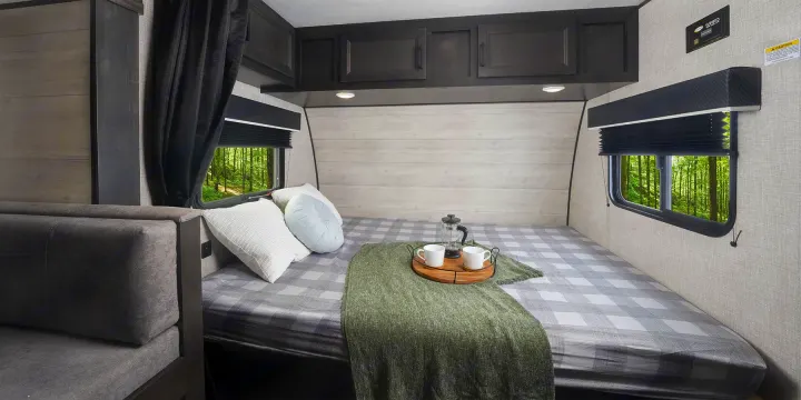 Cozy RV rental bedroom featuring a plaid bed, overhead cabinets, and scenic forest views from the windows.. Jayco slx 2023