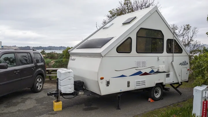 A white A-frame rv rental camper parked at a scenic campsite next to a dark SUV under a cloudy sky.. Chalet Chalet Popup Trailer 2006