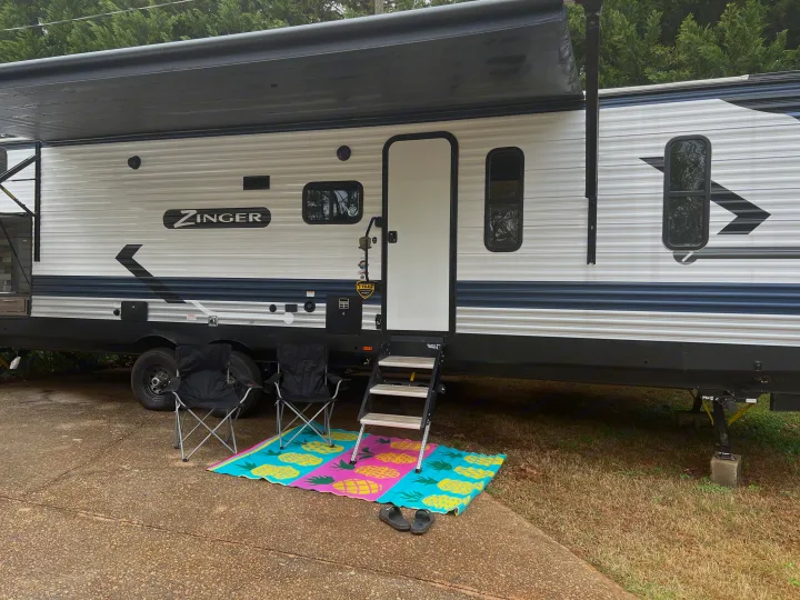 A Zinger travel trailer RV rental with an extended awning provides a cozy outdoor setup: chairs, pineapple mat, and steps.. Crossroads RV Zinger 2022