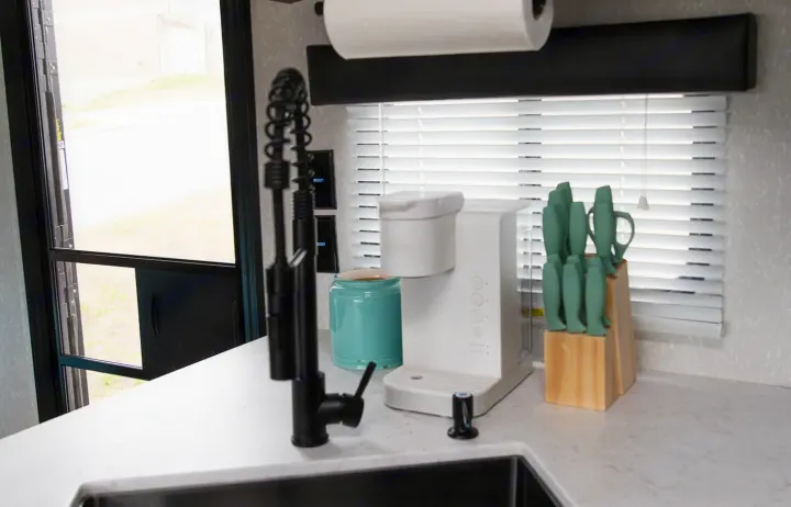 Inside an RV rental: a modern kitchen counter features a white coffee maker, sink, and green knife set by a window.. Forest River Cherokee Grey Wolf 2022