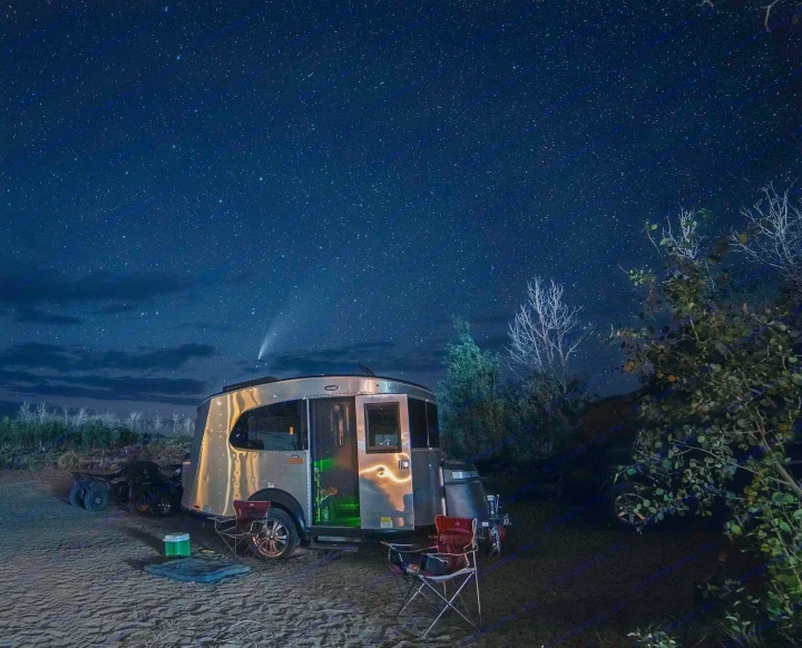 A polished RV rental sits on sandy ground with chairs set up, bathed in light from a spectacular starry night sky and a bright comet.. Airstream Basecamp 2020