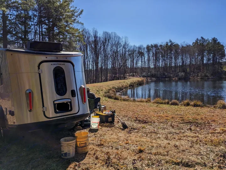 An Airstream Basecamp RV rental parked beside a calm lake with a tree-lined shore, offering a tranquil camping experience.. Airstream Basecamp 2020