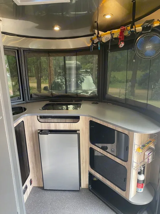 This RV rental's modern kitchen features a compact fridge, counter space, organized storage, and large windows with a forest view.. Airstream Basecamp 2020