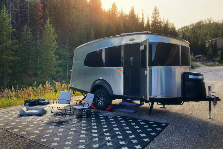 This silver camper trailer RV rental is parked at a mountain campsite with outdoor seating & a rug under golden hour light.. Airstream Basecamp 2020