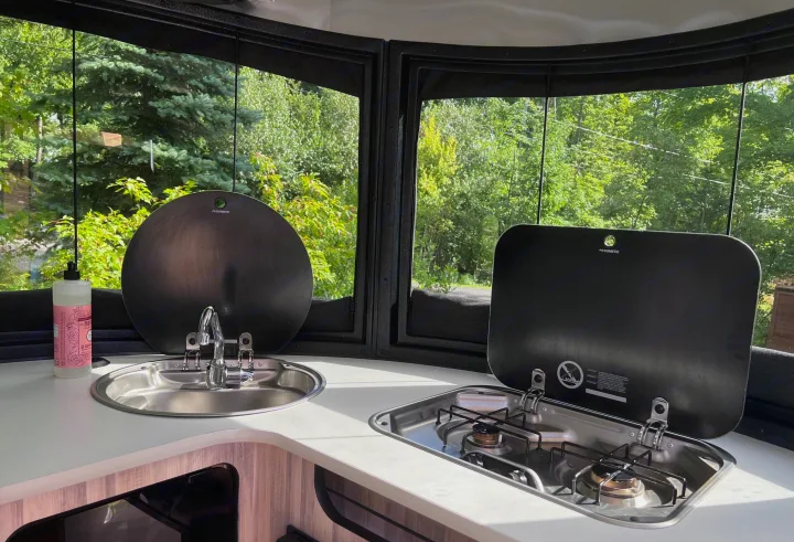 Compact RV rental kitchen featuring a covered sink & two-burner stove. Large windows provide scenic views of lush green trees.. Airstream Basecamp 2020