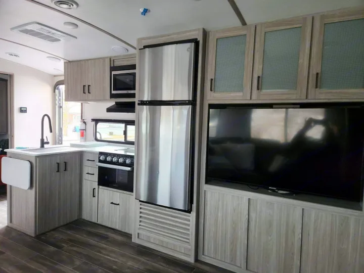 Modern RV rental interior: well-appointed kitchen with stainless appliances, sink, stove, fridge, and entertainment center with TV.. Keystone RV Passport GT 2022