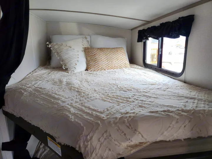 Comfortable RV rental bed with textured white bedding and decorative pillows, offering a cozy retreat.. Keystone RV Passport GT 2022