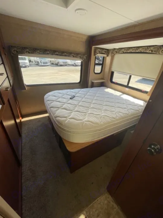 RV rental bedroom with white mattress, windows showing exterior, dark wood cabinetry, and carpeted floor.. Forest River Sunseeker 2015