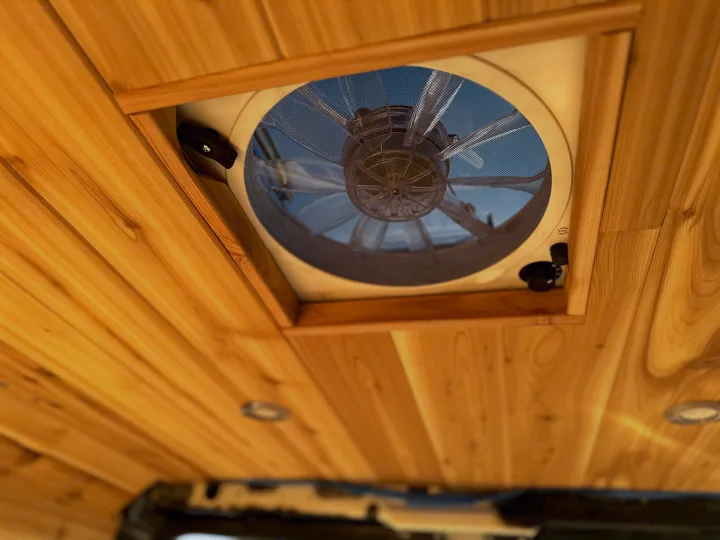 Bright wood-paneled ceiling of an RV rental, featuring a large vent fan with a mesh screen and clear blades looking up at the sky.. Custom Promaster 2021 2021
