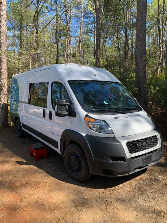 A white "Dave & Matt Vans" camper van (RV rental) is parked on a dirt path in a sunny forest, with a red step stool beside it.. Dodge Sprinter Van 2020