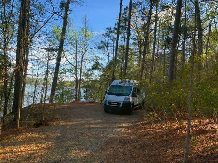 White RV rental van at a peaceful lakeside campsite, nestled among towering pine trees.. Dodge Sprinter Van 2020