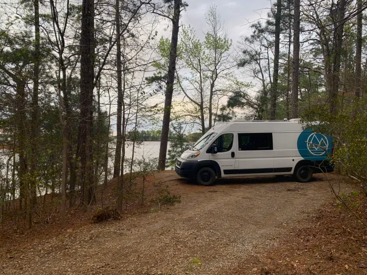 White camper van (RV rental) parked by a lake, surrounded by trees, suggesting a serene outdoor escape.. Dodge Sprinter Van 2020