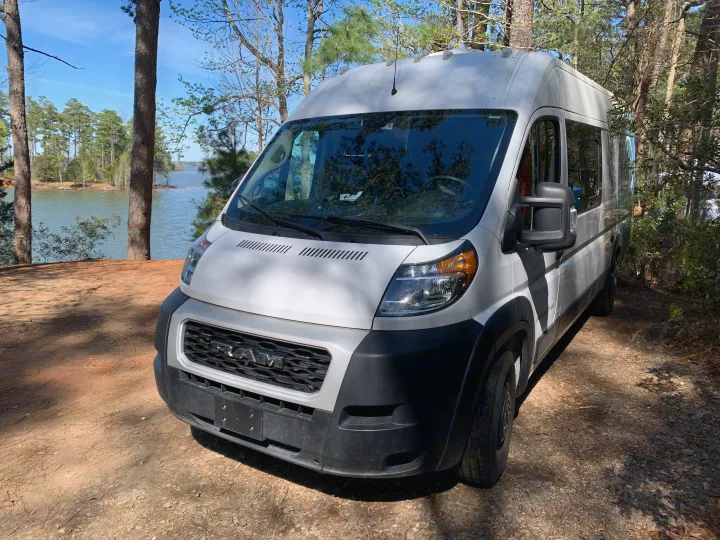A white Ram ProMaster camper van sits by a serene lake, a perfect RV rental for scenic nature trips.. Dodge Sprinter Van 2020