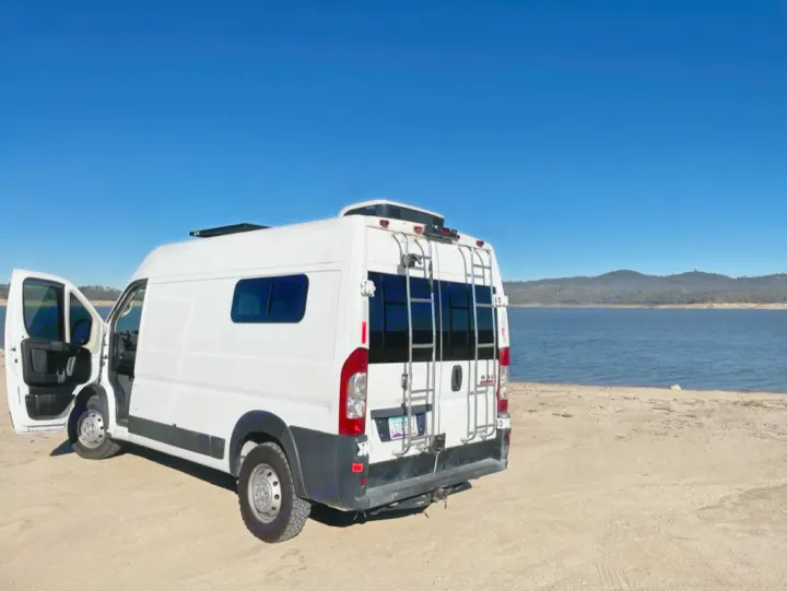 White RV rental camper van parked by a serene lake under a clear blue sky. Perfect nature escape!. Dodge RAM Promaster 2500 2019