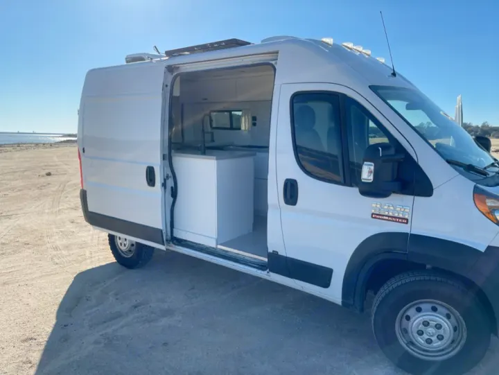 White Ram ProMaster camper van RV rental on a sandy beach. Inside shows a modern kitchen. Perfect for van life travel.. Dodge RAM Promaster 2500 2019