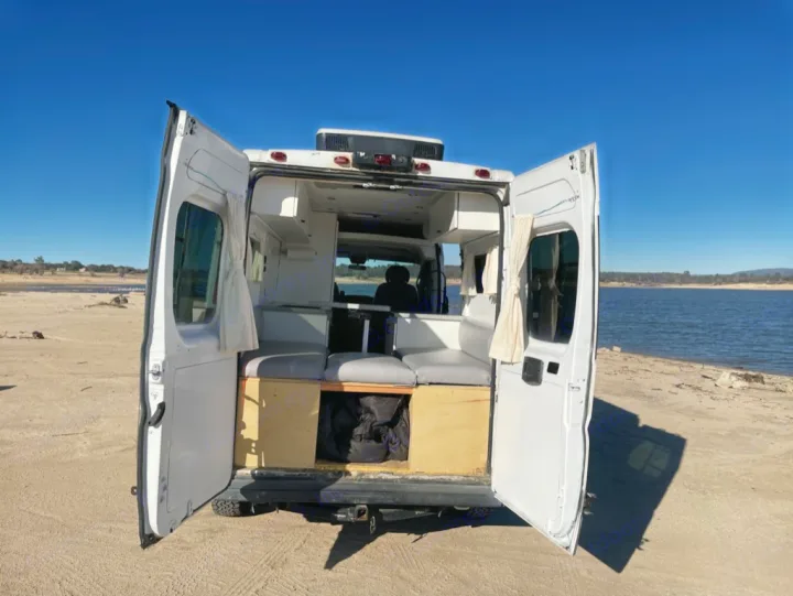 Ready for an **RV rental** adventure! White camper van with open back revealing a cozy interior, parked by a scenic lake.. Dodge RAM Promaster 2500 2019
