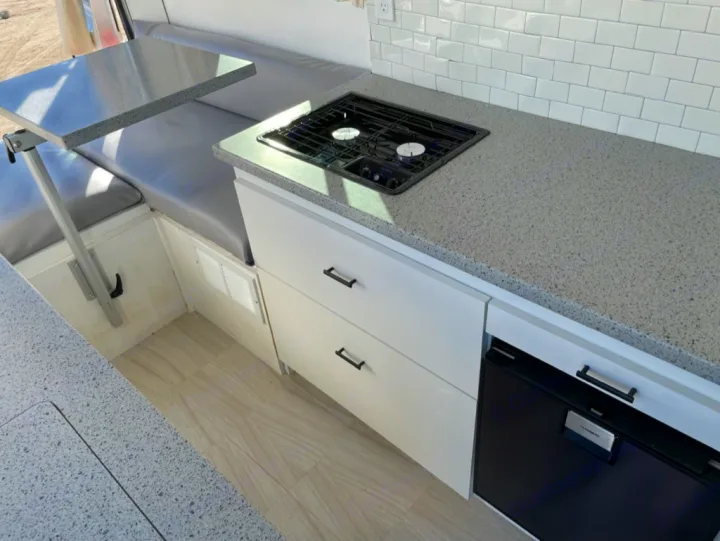 Modern RV rental interior: compact kitchen with gray counters, two-burner stove, fridge, and drop-down dining table.. Dodge RAM Promaster 2500 2019