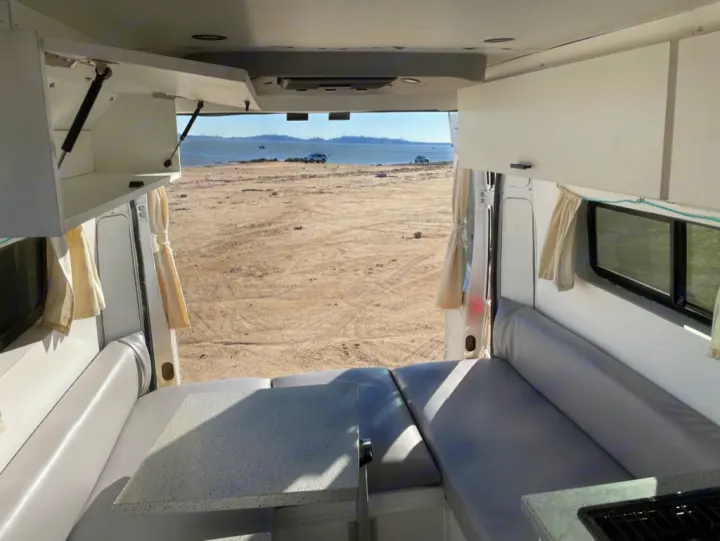 RV rental interior with comfortable seating, a kitchen, and a table, offering a wide view of a sandy landscape by the water.. Dodge RAM Promaster 2500 2019