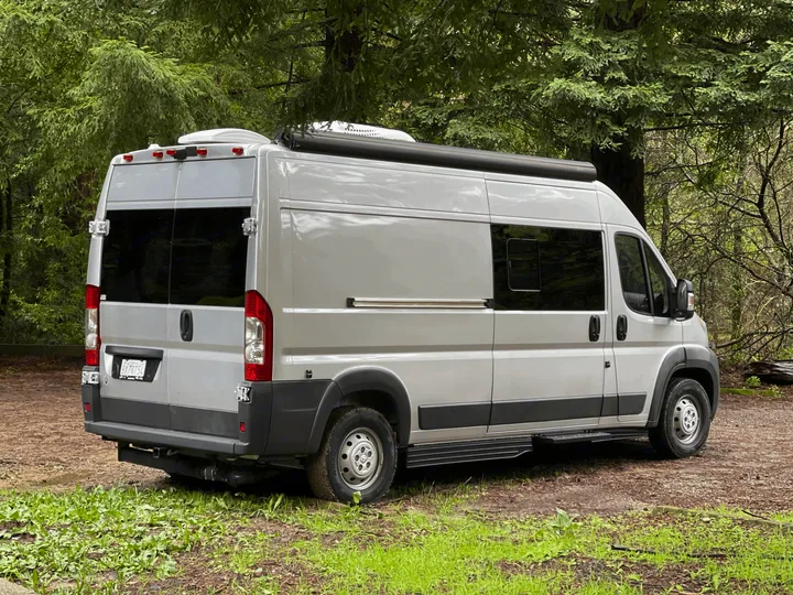 A silver camper van, ideal for RV rental, is parked outdoors. Features include a roof AC unit and awning, surrounded by green trees.. Hymer Sunlight Van 1 2018