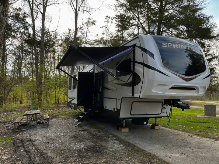 A white Sprinter fifth-wheel RV is set up at a scenic campsite with an extended awning and picnic table. Perfect for an RV rental!. Keystone Sprinter 2023