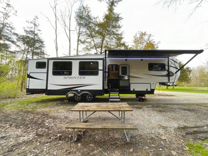 Spacious RV rental at a scenic campsite. Features an extended awning and a wooden picnic table for outdoor relaxation.. Keystone Sprinter 2023