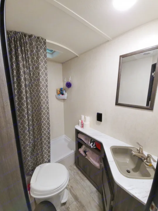 Compact RV rental bathroom featuring a shower with patterned curtain, toilet, vanity with sink, mirror, and storage.. Highland Ridge RV Open Range Conventional OT26BHS 2019