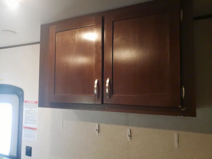Upper wood cabinets with silver handles inside an RV rental. A warning sign and wall hooks are visible below.. Jayco Octane 2019