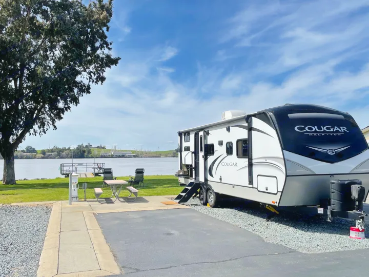 Scenic waterfront RV rental campsite with a white Cougar travel trailer, picnic table, and river views under a blue sky.. Keystone Cougar Half-Ton 2022