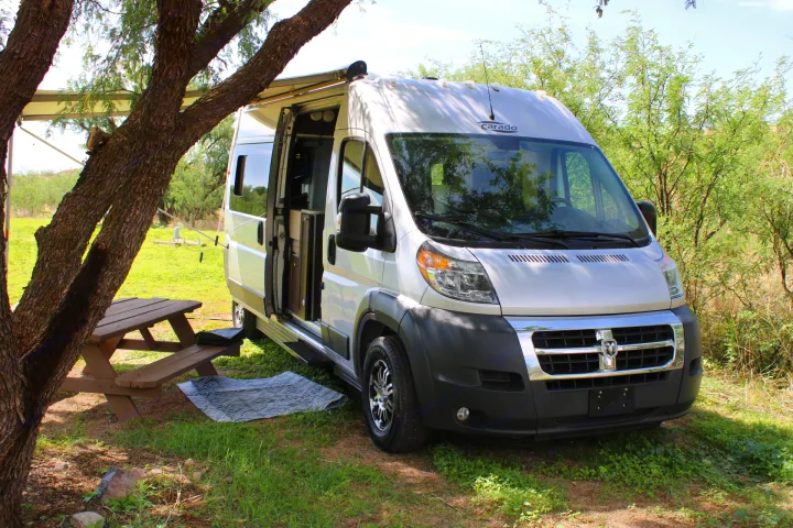 A white Carado camper van, ideal for rv rental adventures, parked under a tree with an awning & picnic table at a campsite.. Carado Carado Class B 2018