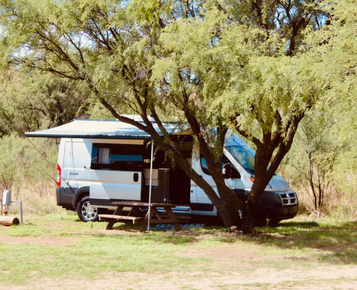 A white RV rental campervan with an extended awning and picnic table sits shaded by trees at a grassy campsite.. Carado Carado Class B 2018