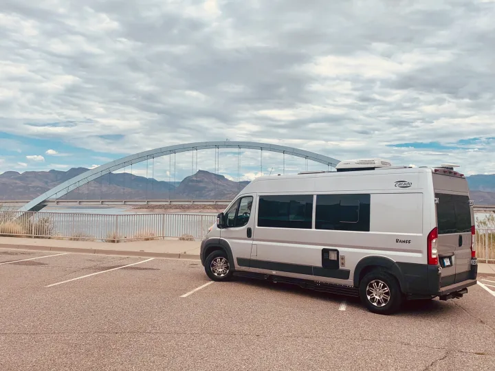 Silver Carado Banff RV rental parked by a scenic arch bridge over water, with mountains in the background under a cloudy sky.. Carado Carado Class B 2018
