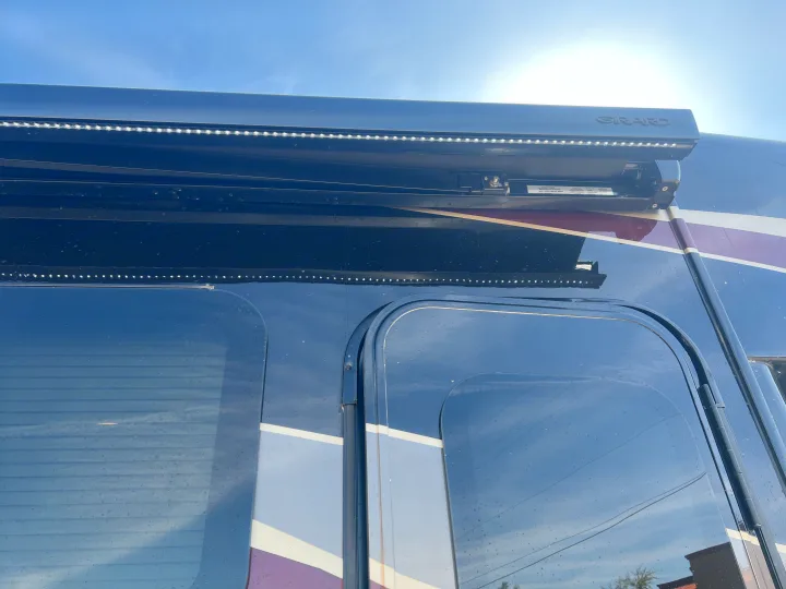 An RV rental's side with a "GIRARD" awning and integrated LED strip lighting. Dark windows with blinds and decorative stripes are also seen.. Entegra Coach Qwest 2020