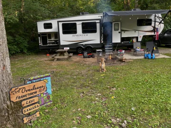 An RV rental at a family campsite, featuring a dog, a child, and a smoky campfire. A personalized sign is on a tree.. Keystone RV Sprinter 2021