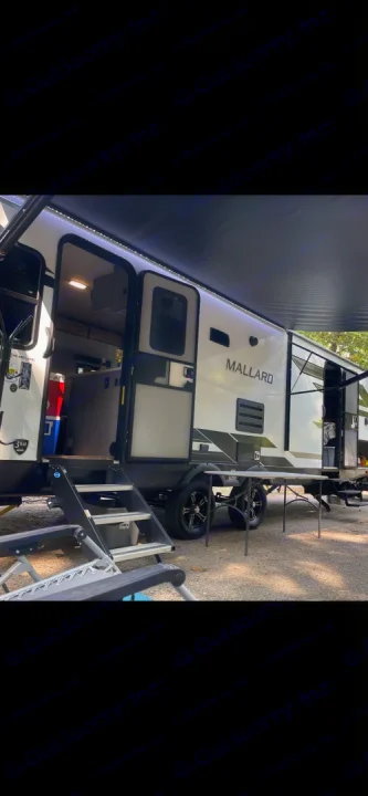 A white and black Mallard **RV rental** is set up at a shaded campsite, with an extended awning, open doors, and outdoor steps.. Heartland RVs Mallard 2022